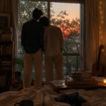 Two young men standing together by a large apartment window at sunrise inside a warm, cozy room filled with books, soft lights, and artistic details.